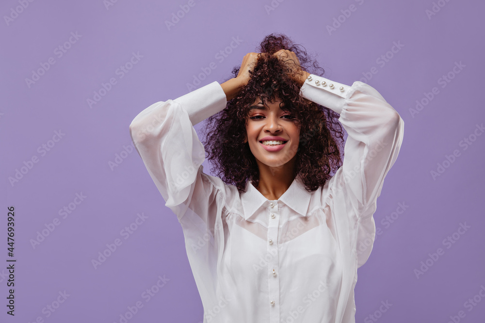 Pretty brunette curly woman in white blouse ruffles hair. Attractive dark-skinned lady smiles and looks into camera on purple background.
