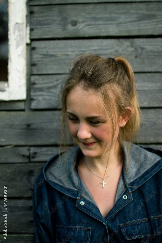 Portrait  of a young embarrassed fair-haired white girl of European appearance at sunset on the background of a wooden wall made of gray boards