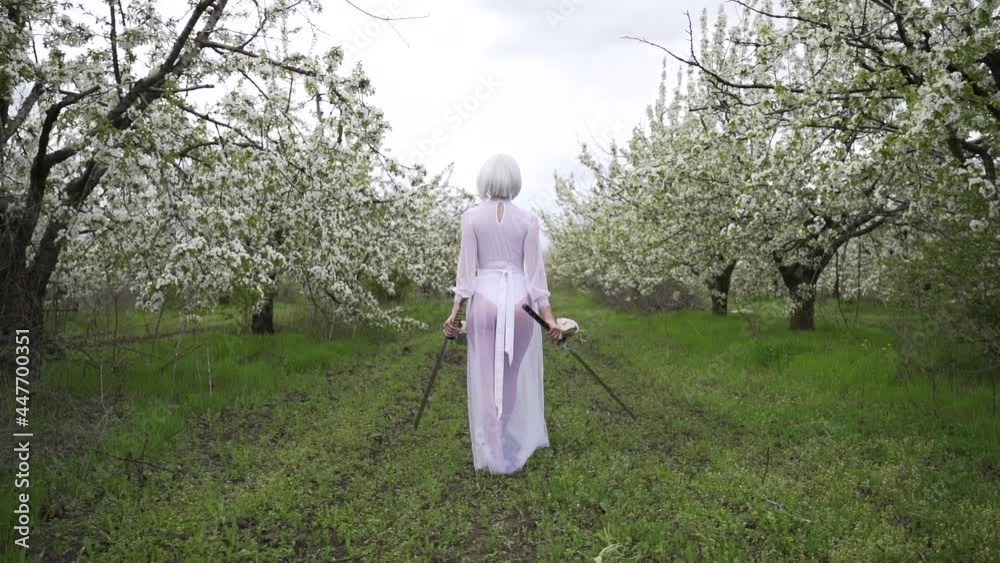 Young blonde in white transparent dress with arms in her hands walks through blooming garden in spring Young blonde in white transparent dress with arms in her hands walks through blooming garden in spring