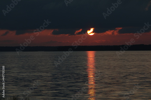 Natural landscape of sunset black red sky with big golden burning ball of sun in dark cloud and glowing shimmering sun path on water surface