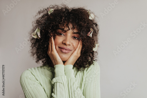 Brunette brown-eyed woman in green top smiles and looks into camera on white background. Young curly lady with flowers in hair poses.