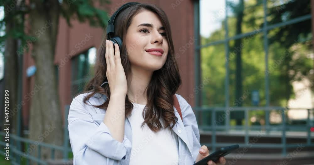 Full length view of the caucasian student woman wearing headphones walking through the street and listening music during the break. Free time concept Full length view of the caucasian student woman wearing headphones walking through the street and listening music during the break. Free time concept