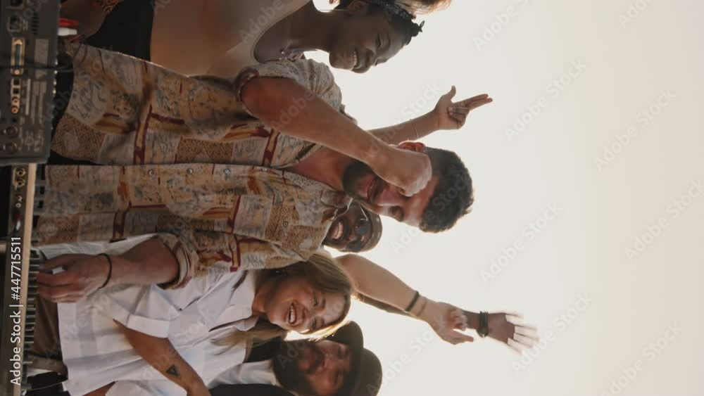 Handheld vertical shot of group of young people dancing and having fun outside on nice summer evening while bearded male DJ playing music