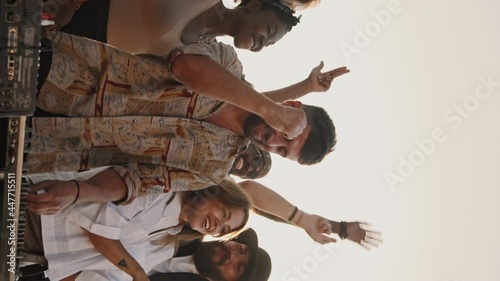 Handheld vertical shot of group of young people dancing and having fun outside on nice summer evening while bearded male DJ playing music photo