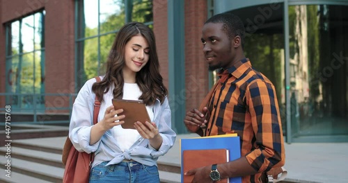 Waist up portrait of the attractive diverse students standing near the university and looking at the tablet computer while discussing something. Education and knowledge concept photo