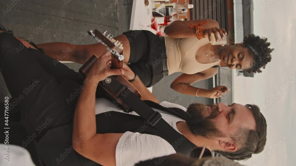 Handheld vertical shot of happy bearded man smiling and playing acoustic guitar for his friends at rooftop party
