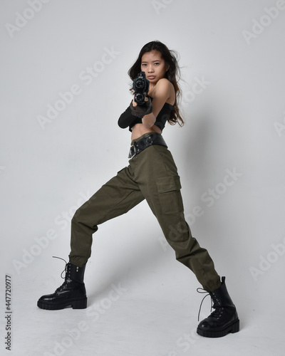 Full length portrait of pretty brunette, asian girl wearing black top and khaki utilitarian army pants and leather boots. Standing pose holding a science fiction gun, isolated agent a light grey studi