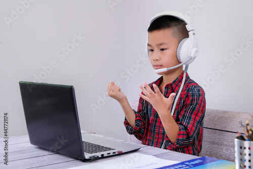 boy sitting in front of laptop screen Study online at home, plug in headphones, hold the answer sheet, show the teacher online, study at home with the teacher, show sign language, count numbers by han