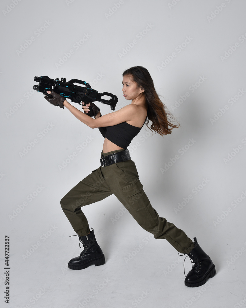 Full length portrait of pretty brunette, asian girl wearing black top and khaki utilitarian army pants and leather boots. Standing pose holding science fiction gun, isolated against studio background Full length portrait of pretty brunette, asian girl wearing black top and khaki utilitarian army pants and leather boots. Standing pose holding science fiction gun, isolated against studio background