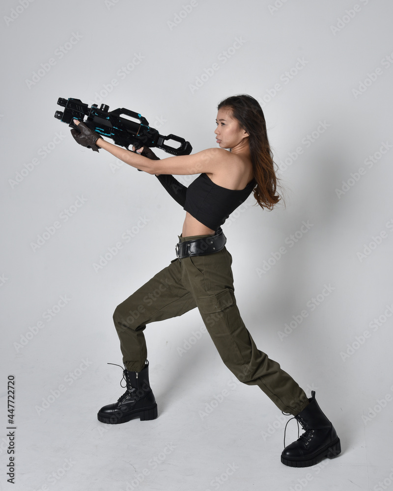 Full length portrait of pretty brunette, asian girl wearing black top and khaki utilitarian army pants and leather boots. Standing pose holding science fiction gun, isolated against studio background Full length portrait of pretty brunette, asian girl wearing black top and khaki utilitarian army pants and leather boots. Standing pose holding science fiction gun, isolated against studio background