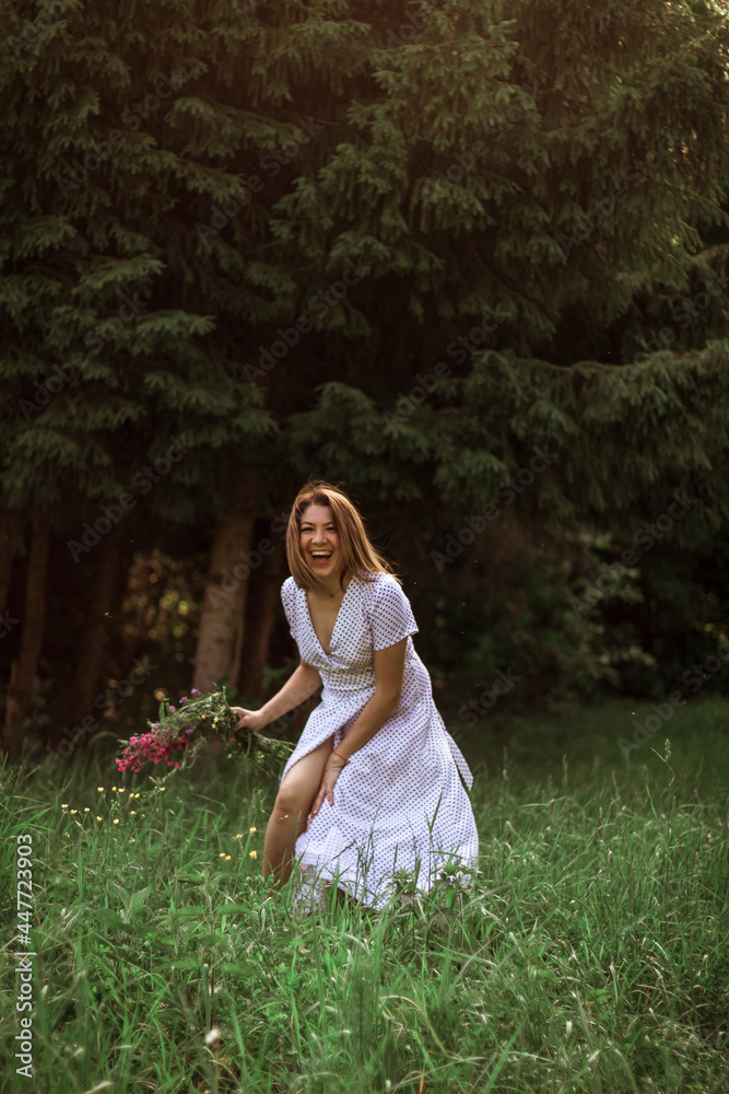 A beautiful happy girl in a long white dress bared her leg, laughing, holding an armful of wild flowers in her hand. Live photo. Selective focus A beautiful happy girl in a long white dress bared her leg, laughing, holding an armful of wild flowers in her hand. Live photo. Selective focus