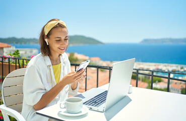 Young elegant woman wearing wireless headphone looking at smartphone with smile while sitting at balcony with seaview on resort. girl watching video, chatting in social network