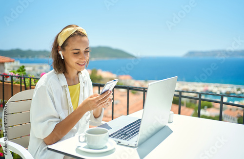 Young elegant woman wearing wireless headphone looking at smartphone with smile while sitting at balcony with seaview on resort. girl watching video, chatting in social network