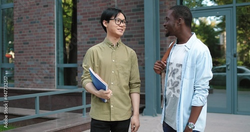 Waist up portrait view of the asian man chatting with his multiracial male classmate while standing with each other at the street near the college during the break. They meeting their female friend photo
