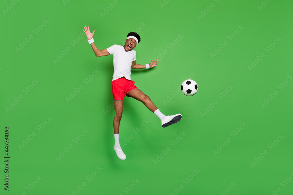 Fototapeta premium Full length photo of strong excited dark skin guy dressed sport outfit jumping playing football isolated green color background