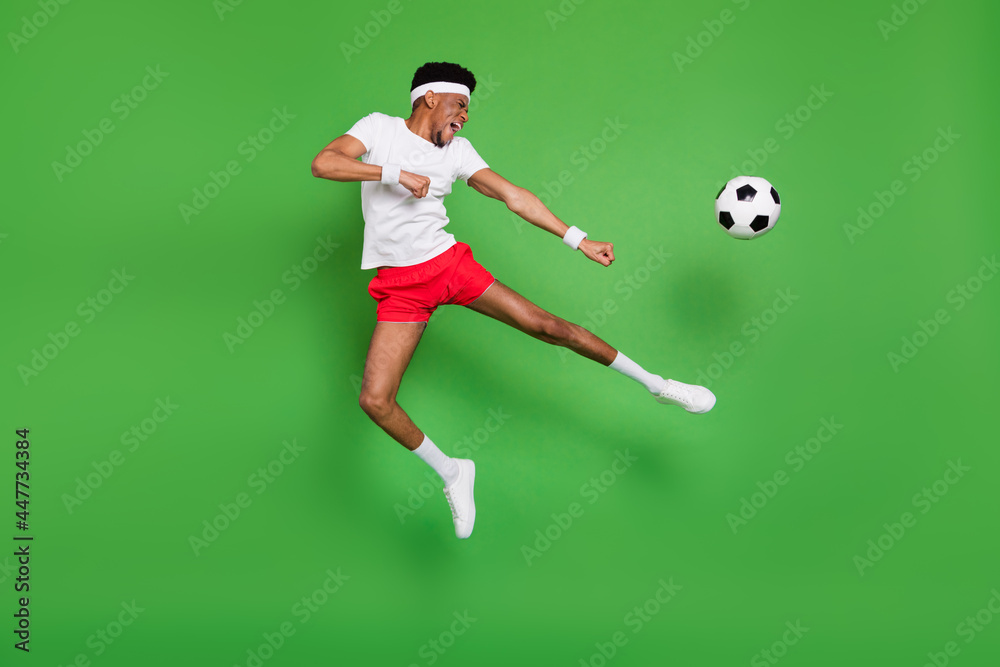 Full length photo of funky strong dark skin guy dressed sport outfit jumping playing football isolated green color background