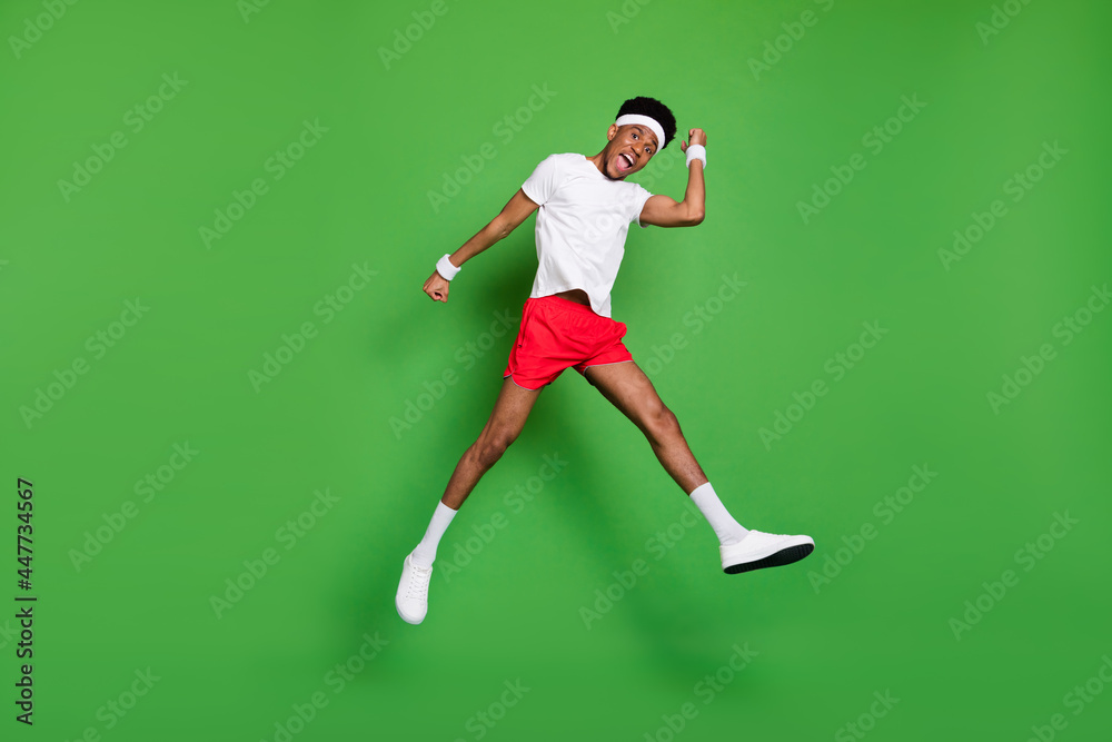 Full length photo of impressed excited dark skin man wear white t-shirt rising fists jumping high isolated green color background Full length photo of impressed excited dark skin man wear white t-shirt rising fists jumping high isolated green color background