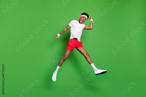 Full length photo of impressed excited dark skin man wear white t-shirt rising fists jumping high isolated green color background