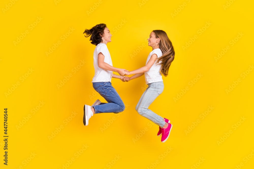 Full length body size view of two nice cheerful carefree kids jumping holding hands isolated over bright yellow color background Full length body size view of two nice cheerful carefree kids jumping holding hands isolated over bright yellow color background