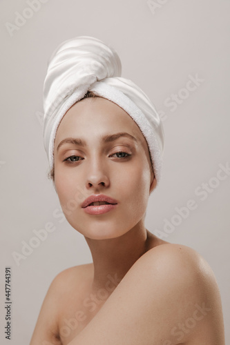 Close-up portrait of attractive young woman in white towel wrapped hair, with pure pale skin, looking into camera against light background. Healthcare and wellness concept