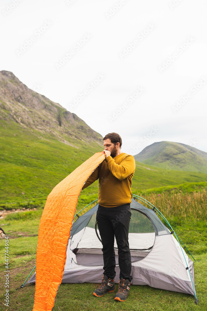Young and attractive man inflating a mattress next to his tent to do solo camping in Scotland. Equipment to travel during vacation