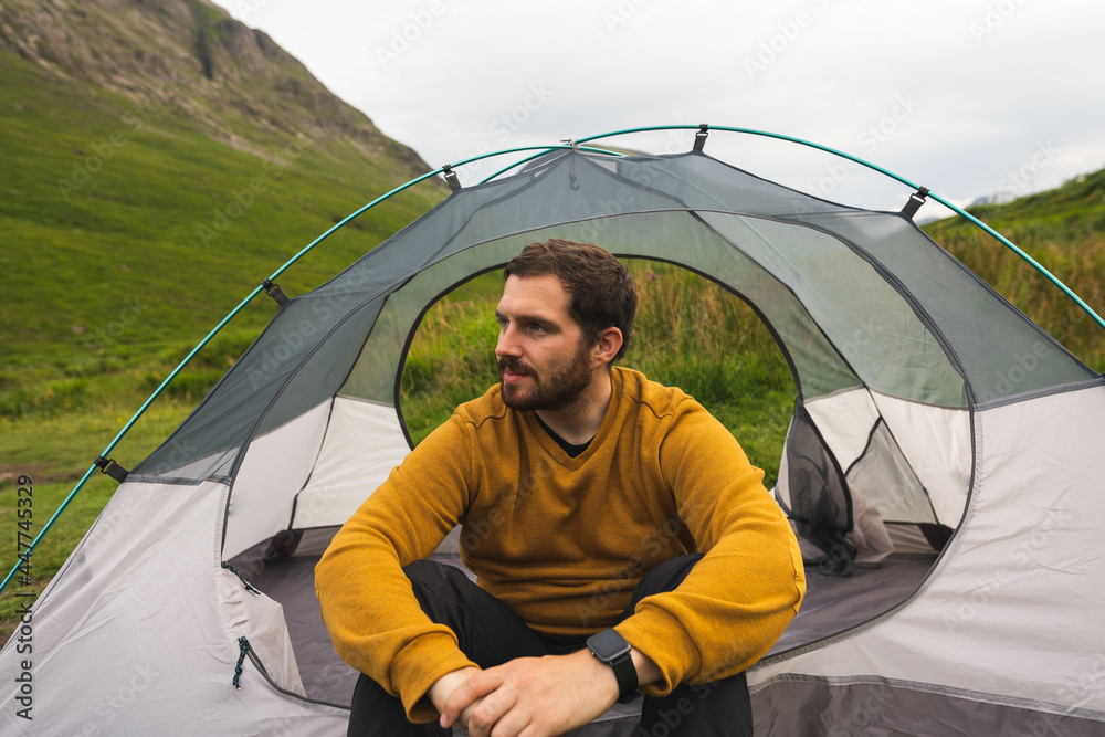 Young and attractive man sitting on his tent set up outdoors enjoying nature in Scotland while doing solo camping