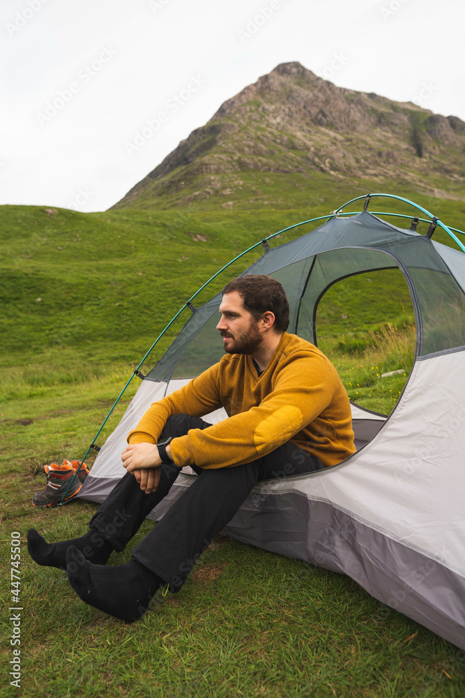 Young and attractive man sitting on his tent set up outdoors enjoying nature in Scotland while doing solo camping
