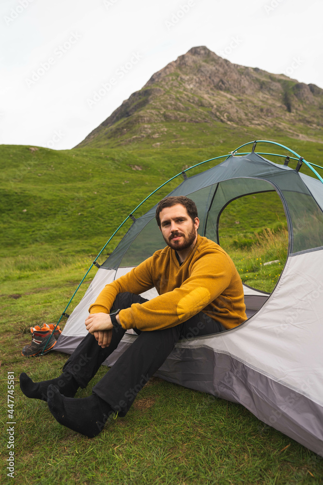 Young and attractive man sitting on his tent set up outdoors enjoying nature in Scotland while doing solo camping