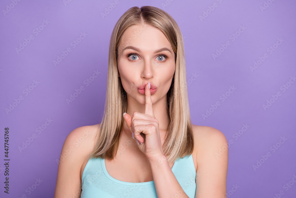 Photo of serious blond young lady finger mouth wear blue top isolated on violet color background Photo of serious blond young lady finger mouth wear blue top isolated on violet color background