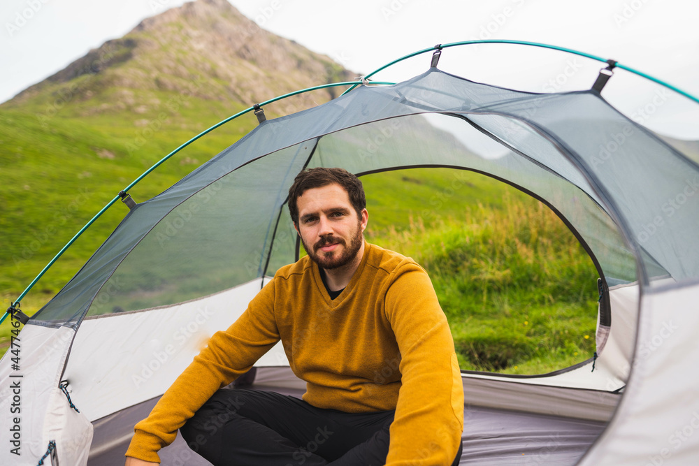 Young and attractive man sitting on his tent set up outdoors enjoying nature in Scotland while doing solo camping