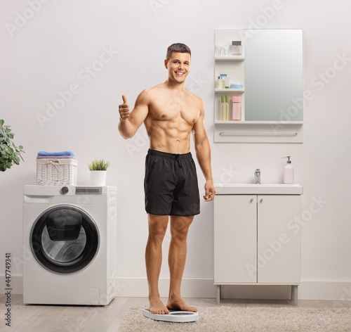 Young smiling shirtless man standing on a weigth scale in a bathroom and showing thumbs up