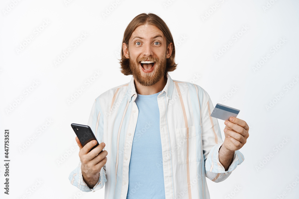 Excited blond man smiling, paying for online shopping, holding credit card with mobile phone, purchase something on sale, standing against white background Excited blond man smiling, paying for online shopping, holding credit card with mobile phone, purchase something on sale, standing against white background