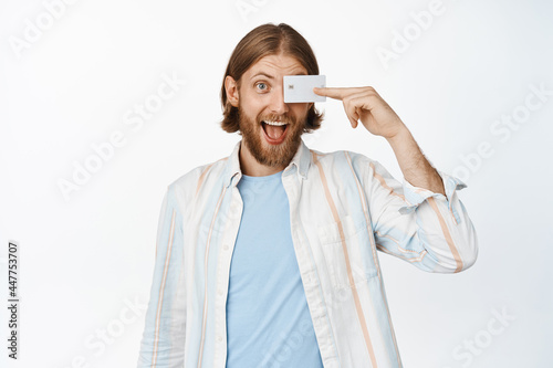 Portrait of excited handsome guy looking happy, holding credit discount card over eye, standing against white background. Shopping and money concept