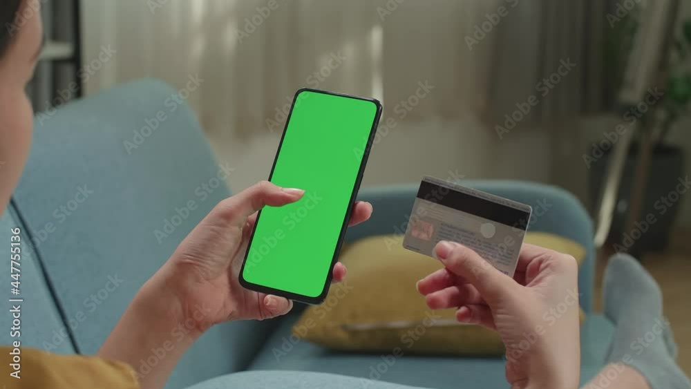 Asian Woman Sitting On Couch In Living Room Using Smartphone With Mock Up Green Screen Display
