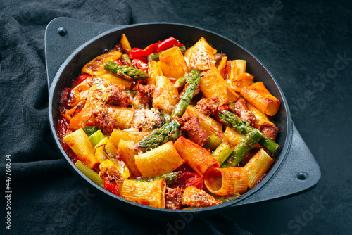 Traditional roasted Italian pasta mezzi paccheri rigati con salsiccia sausage with green asparagus tips in tomato sauce served as close-up in cast iron skillet pan in matte flat cinematic modern look