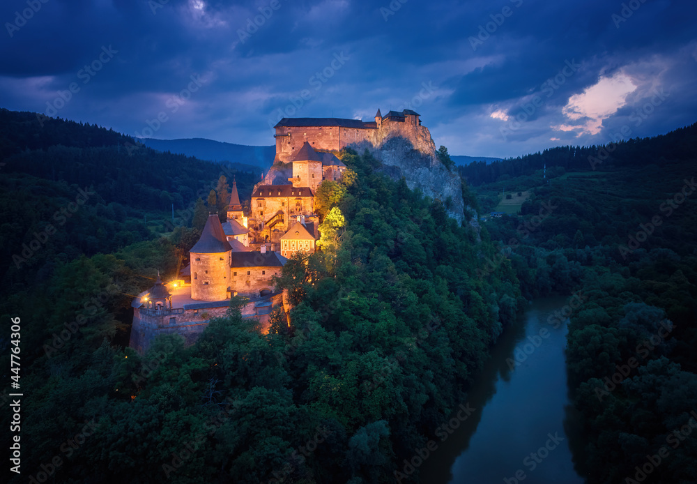 Aerial, dramatic view of Orava Castle situated on a high rock above Orava river, illuminated by orange lamps against dark blue background. Orava, Slovakia.