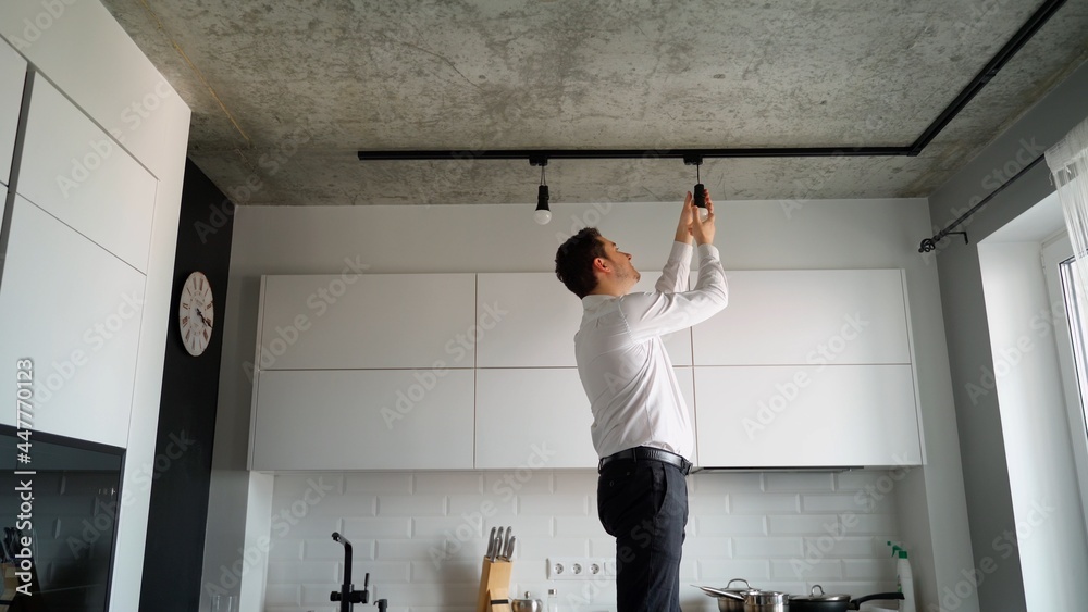 Man of Caucasian ethnicity unscrews a light bulb from the base of modern track lighting of a kitchen at home. He looks like a professional and is neatly dressed in a shirt and trousers
