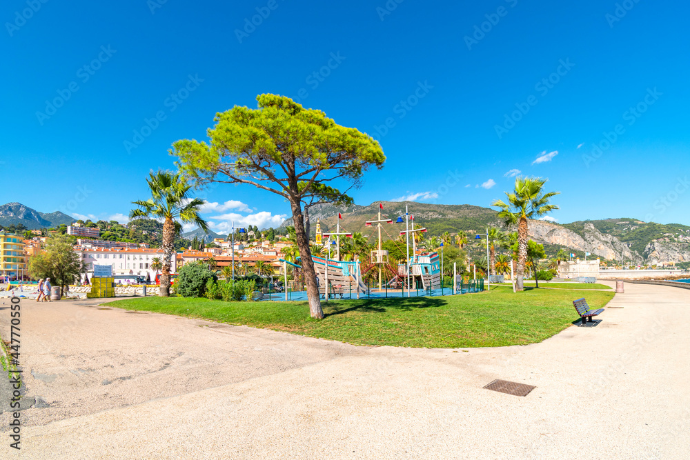 A sunny summer day on the French Riviera as they relax near the sea at Fossan Beach Park in Menton, France, with the mountains and city in view behind.