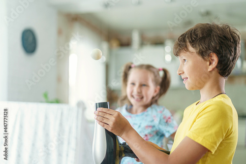 Curious boy and girl conducting a physical experiment called floating ball with a hairdryer and a plastic pink ball at home in the kitchen. Children are happy and laughing and jumping merrily