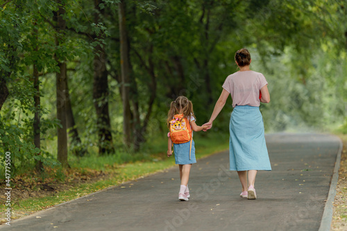 friendly family goes to kindergarten. woman morally supports daughter, holding hands, encourages child. mother accompanies schoolgirl to school. happy little girl with caring mother looks from back photo