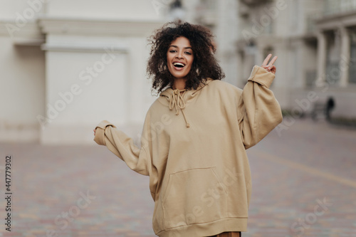 African curly woman smiles and shows peace sign outside. Attractive dark-skinned lady in oversized beige hoodie walks outdoors. photo