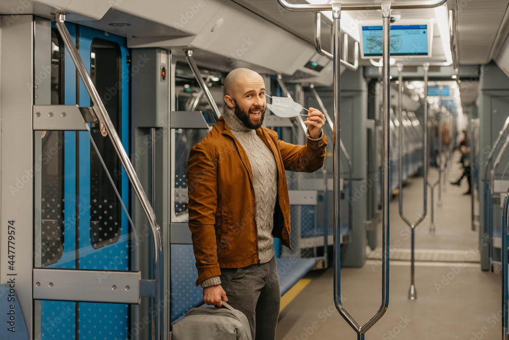 A man with a beard is taking off a medical face mask and smiling on a train.