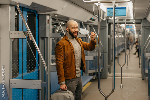 A man with a beard is taking off a medical face mask and smiling on a train.