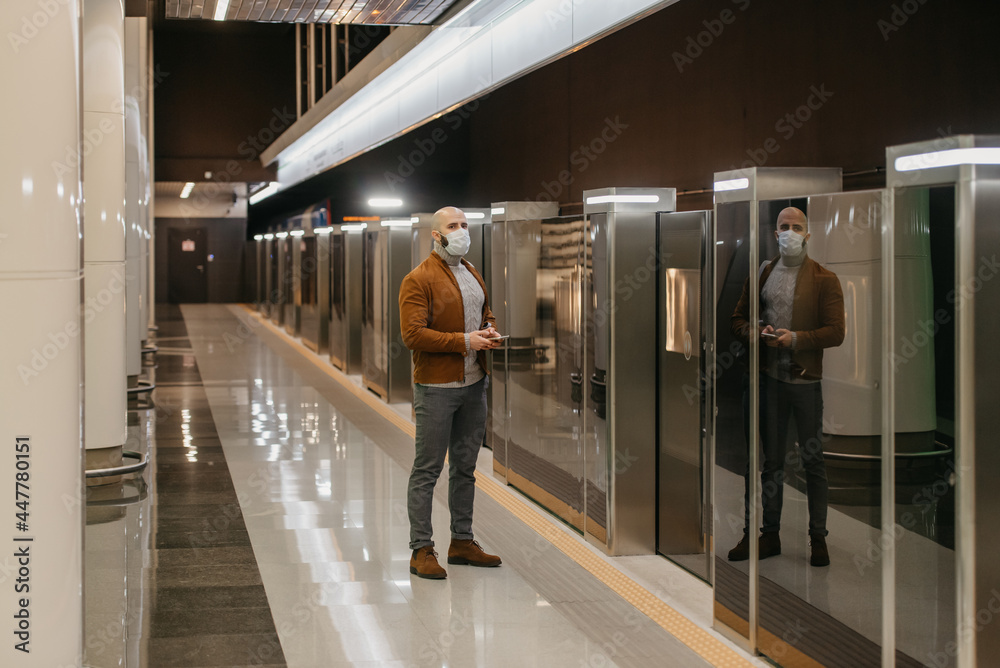 Fototapeta premium A man in a face mask is holding a smartphone while waiting for a subway train.