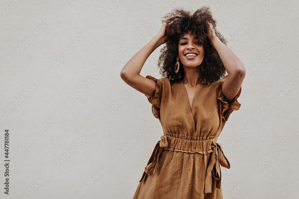 Fototapeta premium Attractive lady in brown dress ruffles hair outside. Charming dark-skinned curly woman in stylish summer outfit smiles on grey background.