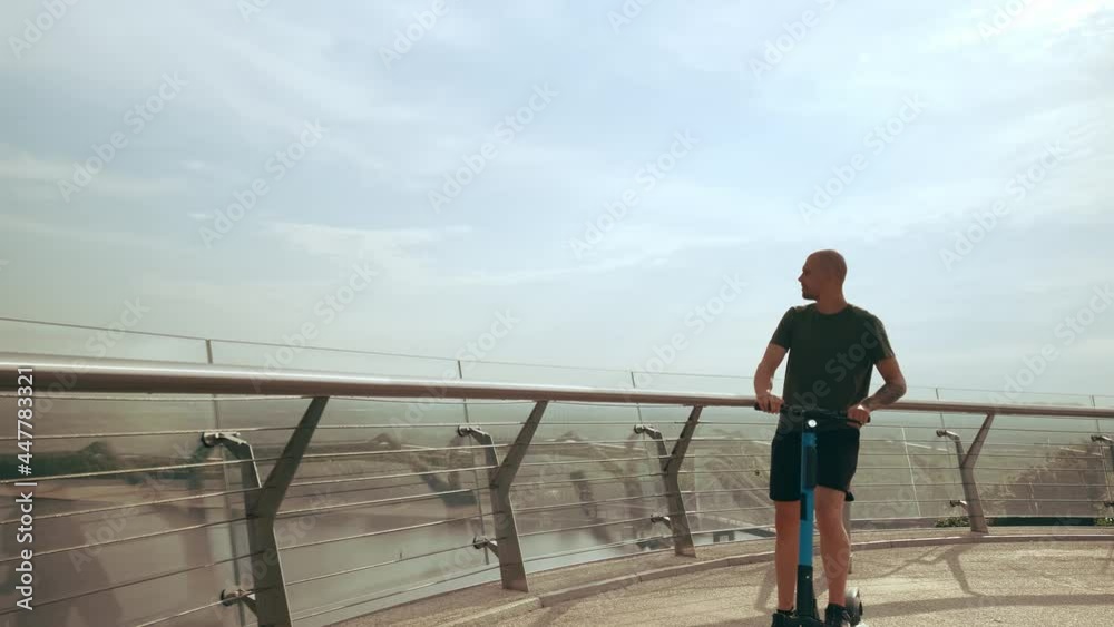 Modern young man rides an electric scooter across the pedestrian bridge overlooking the city. Modern eco transport. High quality 4k footage Modern young man rides an electric scooter across the pedestrian bridge overlooking the city. Modern eco transport. High quality 4k footage