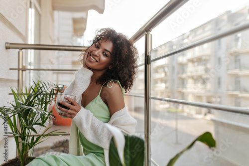 Free Download Charming curly woman drinks coffee on terrace. Young dark-skinned lady in silk green dress and white cardigan smiles. - GFXLoad