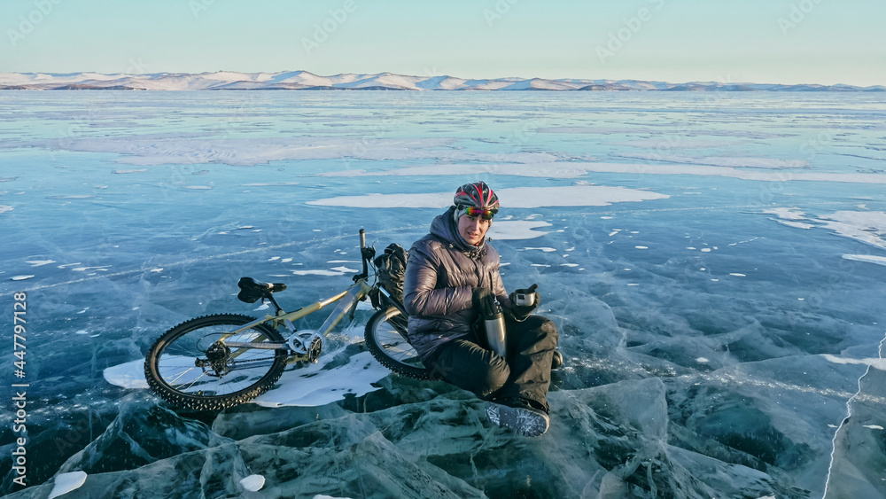 Man sitting near her bike on ice. Athlete cyclist stopped to rest. She sits on the wheel and enjoys the beautiful view of the sunset. The traveler is ride a cycle. Male is riding bike on the ice.