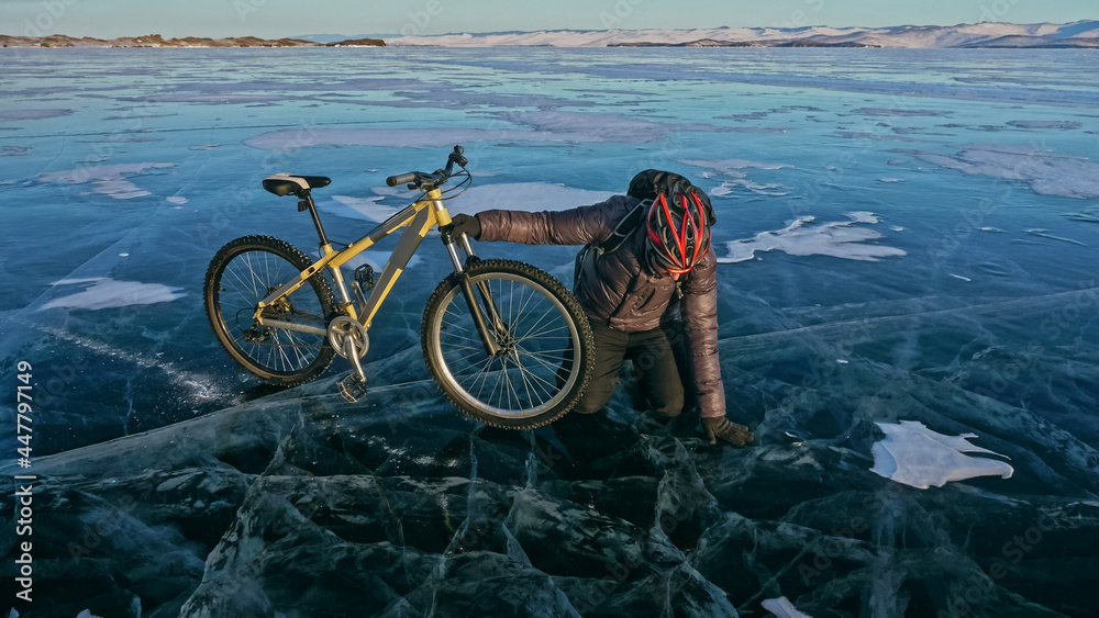 Fototapeta premium Man is riding bicycle on the ice. Ice of the frozen Lake Baikal. Rider is dressed in black down jacket, cycling backpack, helmet. Tires on covered with special spikes. Traveler boy is ride cycle.