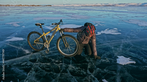 Man is riding bicycle on the ice. Ice of the frozen Lake Baikal. Rider is dressed in black down jacket, cycling backpack, helmet. Tires on covered with special spikes. Traveler boy is ride cycle.